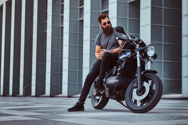 Homme barbu brutal en lunettes de soleil vêtu d'un t-shirt gris et d'un pantalon noir assis sur sa moto rétro sur mesure contre un gratte-ciel.