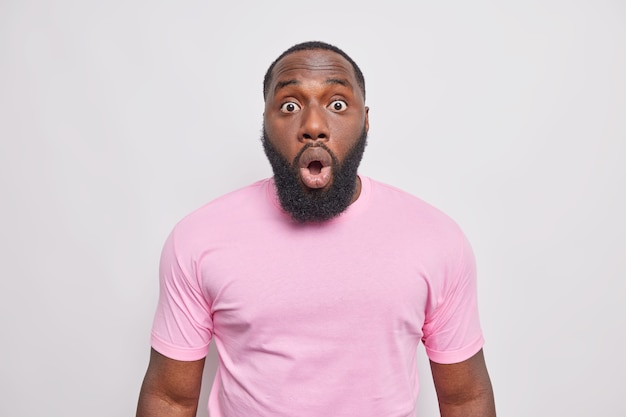 Un homme barbu choqué et inquiet regarde les yeux obsédés à l'avant, ouvre la bouche avec étonnement, réagit à des nouvelles surprenantes vêtus d'un t-shirt rose décontracté isolé sur un mur blanc