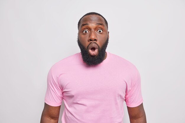 Un homme barbu choqué et inquiet regarde les yeux obsédés à l'avant, ouvre la bouche avec étonnement, réagit à des nouvelles surprenantes vêtus d'un t-shirt rose décontracté isolé sur un mur blanc