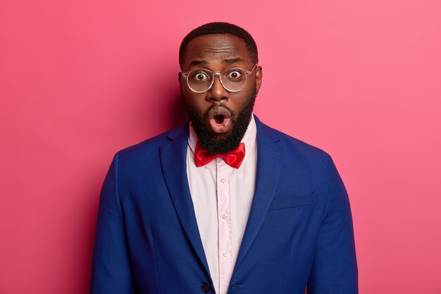 Un homme barbu choqué et inquiet a des soies épaisses, des yeux écarquillés, garde la bouche ouverte, porte des lunettes transparentes, un costume formel avec un nœud papillon