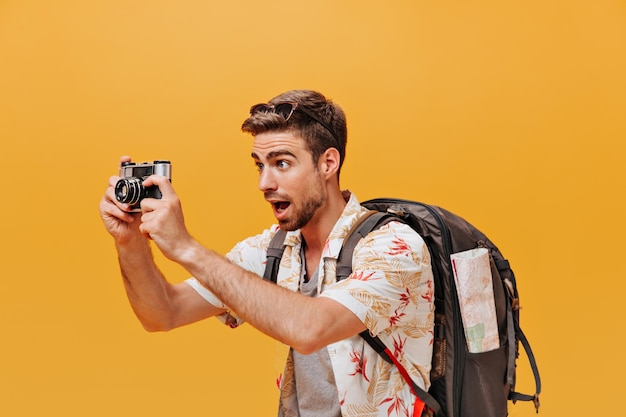 Photo gratuite homme barbu choqué avec des lunettes de soleil en chemise imprimée légère posant avec un grand sac à dos et prenant des photos sur fond isolé