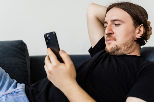 Homme barbu confiant est assis sur le canapé et tapant sur smartphone