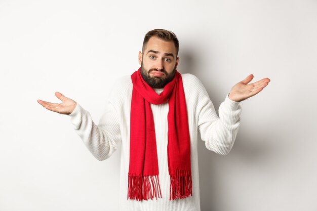 Homme barbu confus haussant les épaules, levant les mains et l'air désemparé, ne sait rien, debout dans un pull et une écharpe de Noël, fond blanc