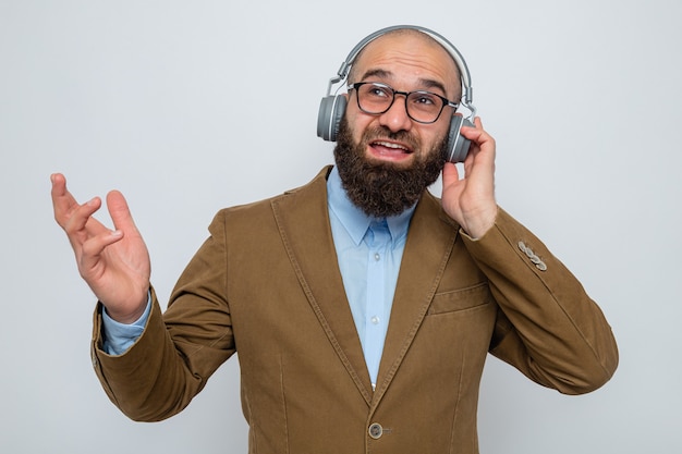 Photo gratuite homme barbu en costume marron portant des lunettes avec un casque à la recherche de sourire heureux et gai debout sur fond blanc