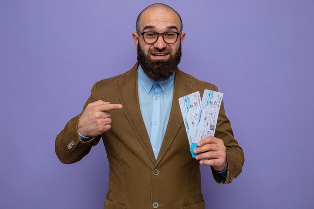 Homme barbu en costume marron portant des lunettes tenant des billets d'avion pointant avec l'index sur eux souriant joyeusement heureux et positif