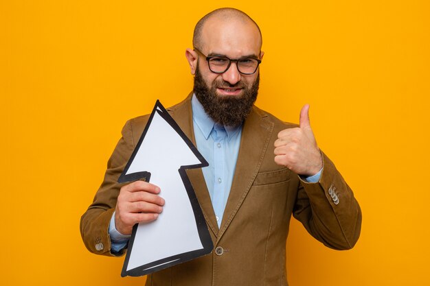 Homme barbu en costume marron portant des lunettes tenant une flèche regardant la caméra souriant joyeusement montrant les pouces vers le haut debout sur fond orange