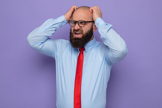 Homme barbu en cravate rouge et chemise bleue portant des lunettes à l'air agacé et frustré de crier et de crier