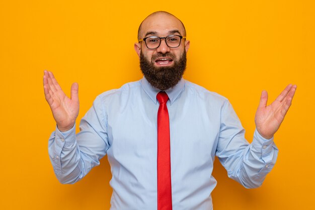 Homme barbu en cravate rouge et chemise bleue portant des lunettes regardant la caméra souriant heureux et positif levant gaiement les mains debout sur fond orange