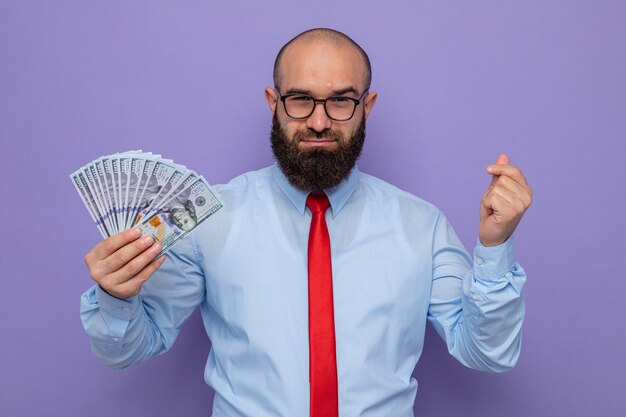 Homme barbu en cravate rouge et chemise bleue portant des lunettes tenant de l'argent en regardant la caméra heureux et confiant souriant faisant un geste d'argent se frottant les doigts debout sur fond violet