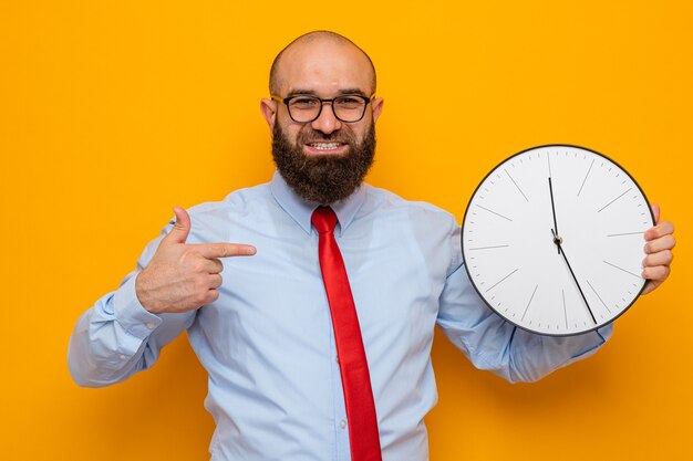 Homme barbu en cravate rouge et chemise bleue portant des lunettes tenant une horloge pointée avec l'index souriant joyeusement