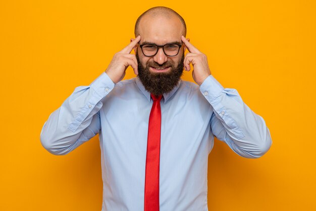 Homme barbu en cravate rouge et chemise portant des lunettes regardant la caméra pointant l'index sur ses tempes en se concentrant sur une tâche debout sur fond orange