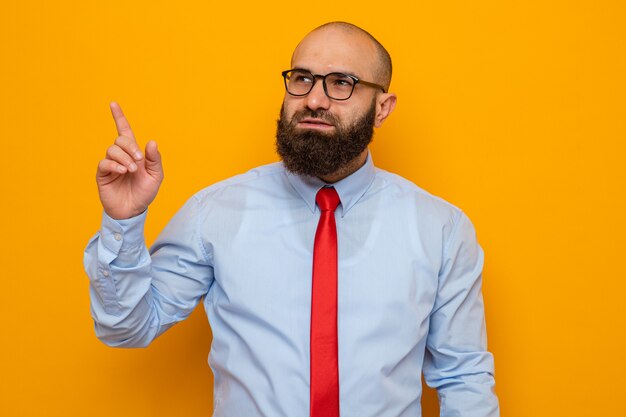 Homme barbu en cravate rouge et chemise portant des lunettes regardant de côté avec un sourire sur un visage intelligent pointant avec l'index vers quelque chose
