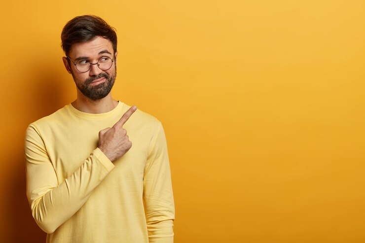 Photo gratuit un homme barbu curieux montre un espace de copie sur le mur jaune, montre des informations importantes, trouve une solution ou une réponse à une question, dit sa suggestion, porte un pull jaune, des lunettes rondes
