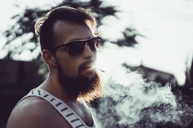 un homme barbu dans les lunettes de soleil fume une cigarette au coucher du soleil, libère une fumée de tabac épais