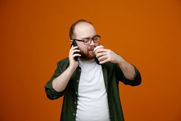 Homme barbu dans des vêtements décontractés portant des lunettes parlant au téléphone mobile tenant une tasse de café à la recherche de confiance heureux et positif debout sur fond orange