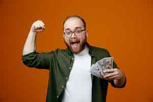 Photo gratuite homme barbu dans des vêtements décontractés portant des lunettes tenant de l'argent serrant le poing heureux et positif se réjouissant de son succès debout sur fond orange