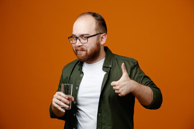 Homme barbu dans des vêtements décontractés portant des lunettes tenant un verre d'eau regardant la caméra heureux et positif montrant le pouce vers le haut souriant confiant debout sur fond orange