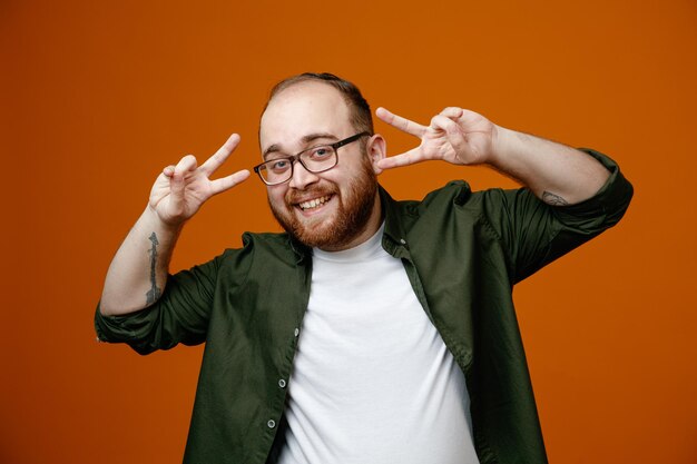 Homme barbu dans des vêtements décontractés regardant la caméra heureux et joyeux montrant des vsigns souriant debout sur fond orange