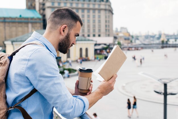 Homme barbu debout près de la balustrade tenant une tasse de café jetable en lisant un livre
