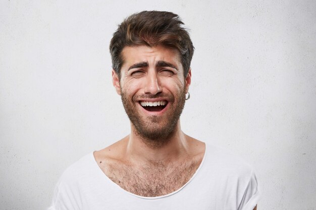 Homme barbu drôle positif avec une coiffure élégante fermant les yeux tout en souriant sincèrement. Joyeux mec attrayant avec une barbe noire fronçant les sourcils de joie