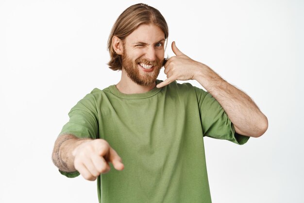 Homme barbu effronté clignant de l'œil montrant appeler mon numéro geste téléphone près de l'oreille pointant du doigt la caméra vous debout sur fond blanc Espace de copie