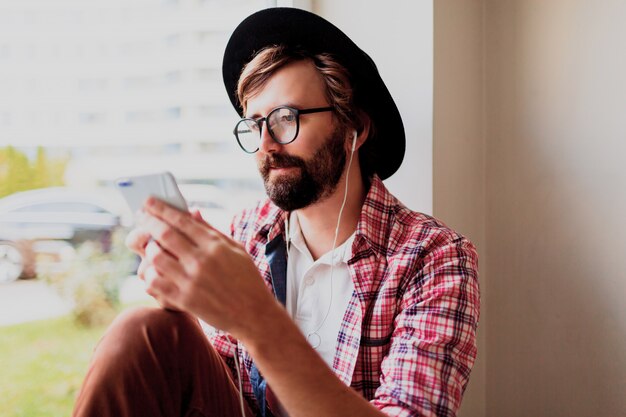 Homme barbu élégant en chemise à carreaux brillants, installant une nouvelle application mobile sur un smartphone et écoutant de la musique. Style hipster.