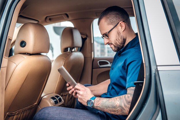 Homme barbu élégant dans des lunettes avec tatouage sur son bras à l'aide d'une tablette PC portable sur un siège arrière d'une voiture.