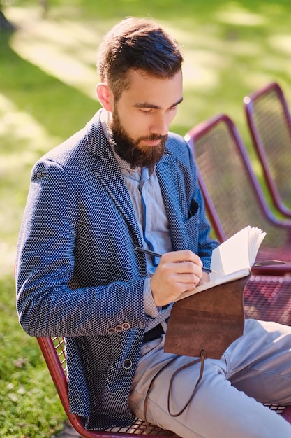 Homme barbu élégant écrivant un message sur un bloc-notes dans un parc.