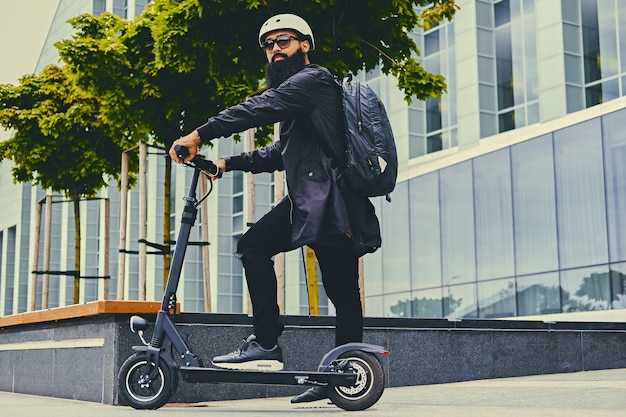 Photo gratuite homme barbu élégant en lunettes de soleil posant sur un scooter électrique sur fond de bâtiment moderne.