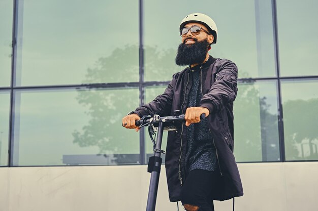 Homme barbu élégant en lunettes de soleil posant sur un scooter électrique sur fond de bâtiment moderne.