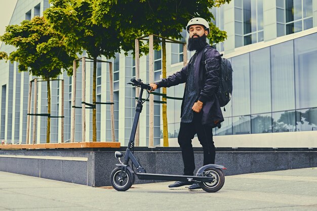 Homme barbu élégant en lunettes de soleil posant sur un scooter électrique sur fond de bâtiment moderne.