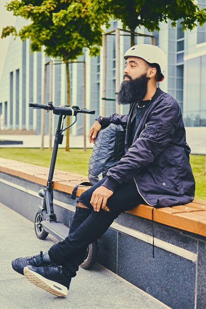 Homme barbu élégant en lunettes de soleil se reposant sur un banc après avoir roulé en scooter électrique au centre-ville.