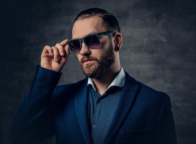 Un homme barbu élégant vêtu d'un costume et de lunettes de soleil sur fond gris foncé.