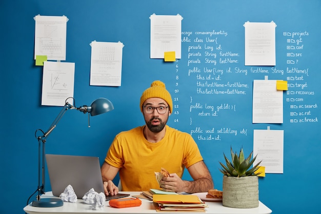 Un homme barbu embarrassé travaille au bureau, a une réaction négative, mange un sandwich, recherche des informations sur un ordinateur portable moderne, une pile de manuels ou des blocs-notes.