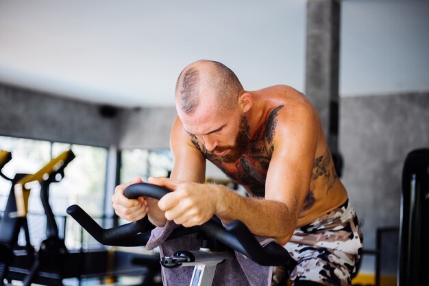 Homme barbu fort musclé tatoué travailler cardio sur vélo dans une salle de sport près de grande fenêtre avec vue sur les arbres à l'extérieur