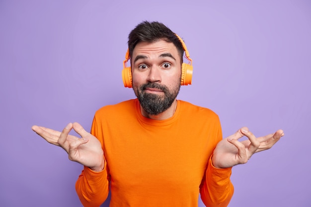 Un homme barbu hésitant et inconscient écarte les mains sur le côté se sent confus a une barbe épaisse a l'air désemparé vêtu d'un pull orange décontracté écoute de la musique avec des écouteurs