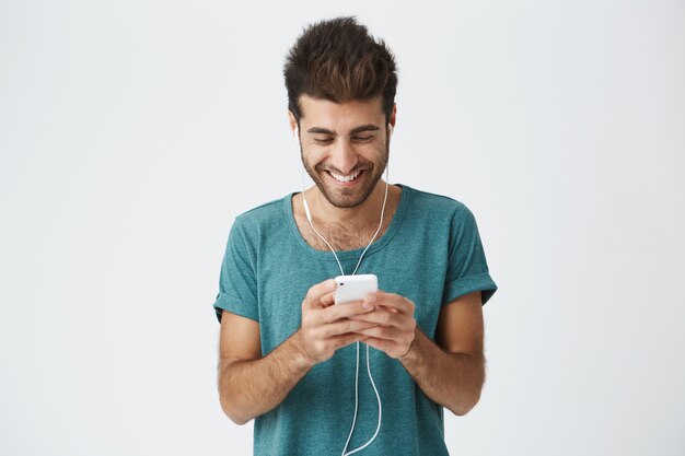 Homme barbu hispanique positif en t-shirt bleu élégant, bonne coiffure et barbe, écoutant de la musique avec des écouteurs et regardant à travers les réseaux sociaux sur son téléphone portable.