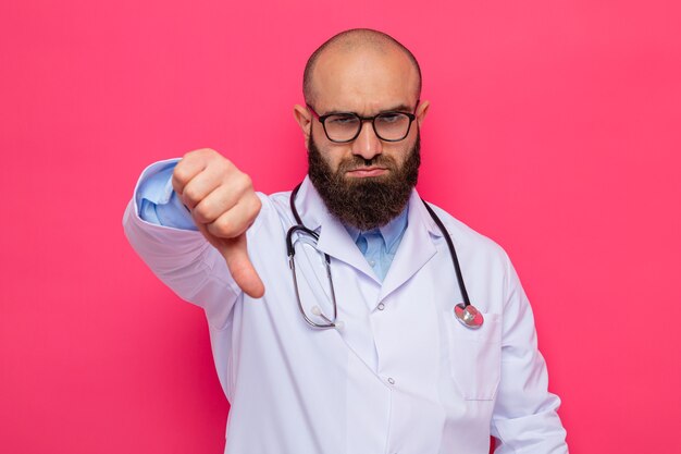 Homme barbu insatisfait médecin en blouse blanche avec stéthoscope autour du cou portant des lunettes regardant avec un visage fronçant les sourcils montrant les pouces vers le bas