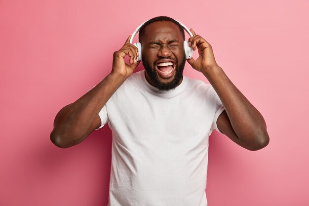 Un homme barbu insouciant détendu et émotionnel écoute la chanson préférée de la liste de lecture, divertit à l'intérieur, porte des écouteurs blancs sur les oreilles, un t-shirt décontracté, pose sur un mur rose.