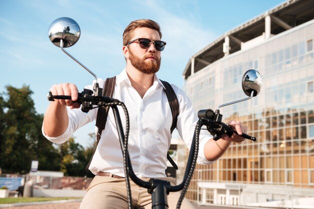 Homme barbu insouciant à lunettes de soleil avec sac à dos monte sur une moto moderne à l'extérieur et à la recherche de suite