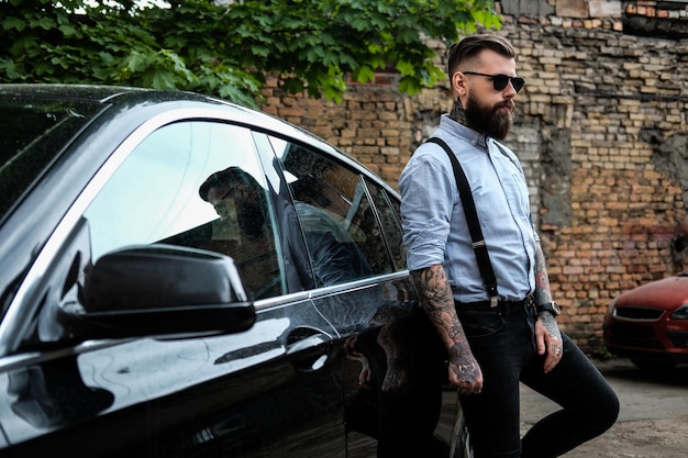 Photo gratuite un homme barbu intelligent s'appuie sur sa voiture. il porte une chemise, des bretelles et des lunettes de soleil. il a des tatouages et une montre sur ses bras. il y a des bidonvilles et des arbres à l'arrière-plan.