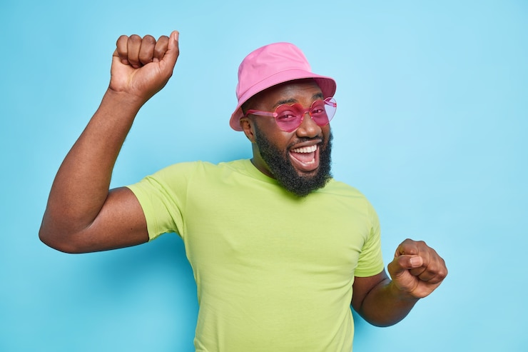 Photo gratuit un homme barbu joyeux danse insouciant garde les bras levés se sent des sourires heureux porte joyeusement des lunettes de soleil de t-shirt vert panama rose a une peau foncée isolée sur un mur bleu