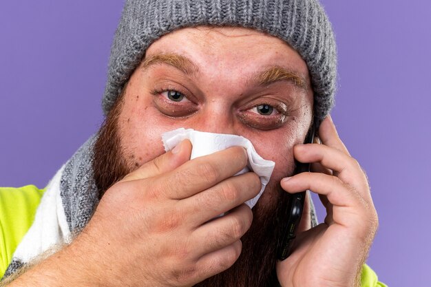 Homme barbu malsain au chapeau et avec une écharpe chaude autour du cou se sentant terriblement souffrant de la grippe parlant au téléphone portable soufflant le nez qui coule éternuant dans un tissu debout sur un mur violet