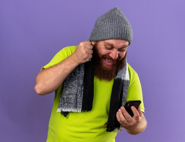 Photo gratuite homme barbu malsain au chapeau et avec une écharpe chaude autour du cou se sentant terriblement souffrant de la grippe avec un téléphone portable qui se déchaîne en criant le poing se tenant debout sur un mur violet