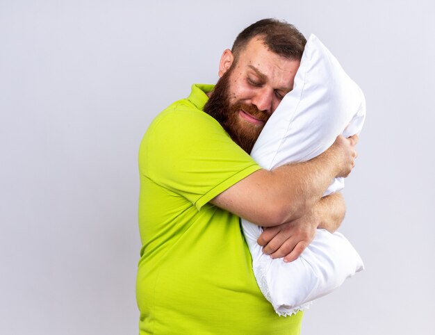 Homme barbu malsain bouleversé en polo jaune se sentant malade tenant un oreiller veut dormir debout sur un mur blanc