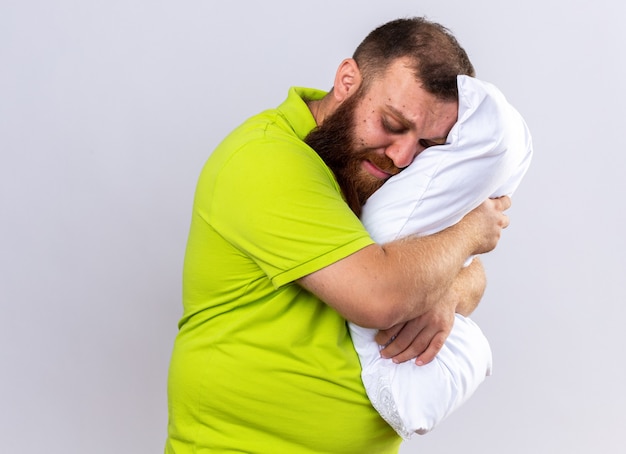 Homme barbu malsain bouleversé en polo jaune se sentant malade tenant un oreiller veut dormir debout sur un mur blanc