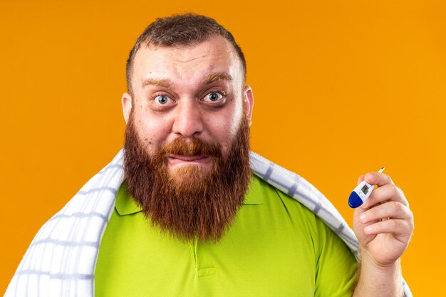 Homme barbu malsain enveloppé dans une couverture se sentant malade souffrant du froid vérifiant la température à l'aide d'un thermomètre l'air inquiet et effrayé debout sur un mur orange