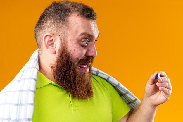 Homme barbu malsain enveloppé dans une couverture se sentant malade souffrant du froid vérifiant la température à l'aide d'un thermomètre stressé et nerveux debout sur un mur orange