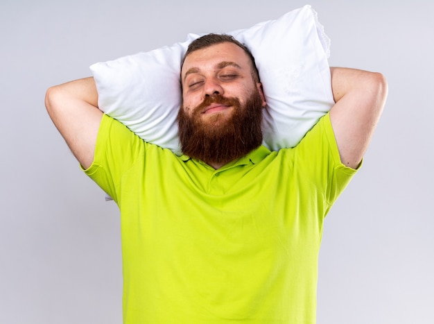 Photo gratuite homme barbu malsain en polo jaune avec oreiller souriant heureux et positif avec les yeux fermés sur un mur blanc