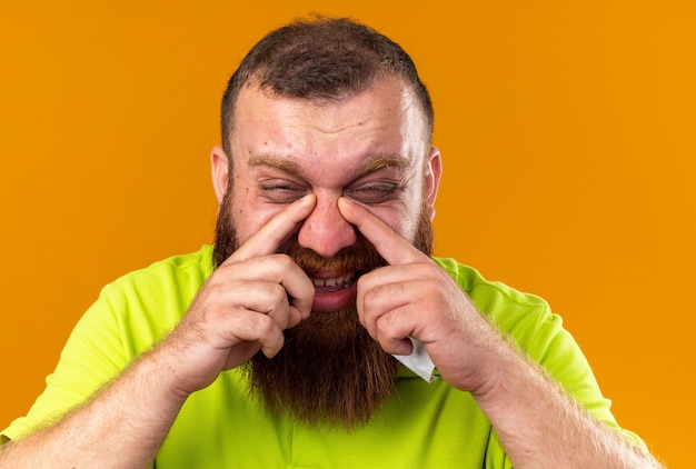 Photo gratuite homme barbu malsain en polo jaune se sentant terriblement souffrant du froid ayant des maux de tête à cause d'un nez bouché debout sur un mur orange